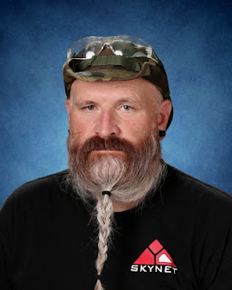 Michael Busse. A white main with a long grey/white beard wearing a camo cowboy hat, a black shirt with the Skynet logo, and safety glasses on top.